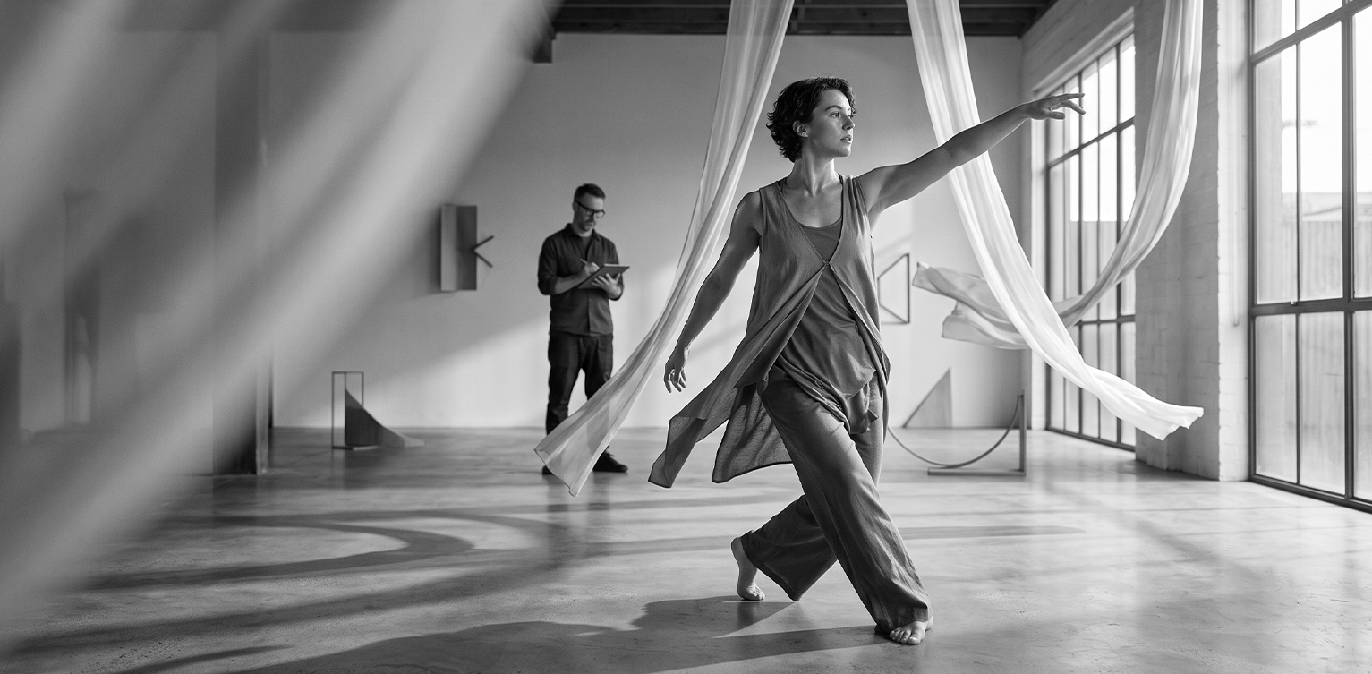 dancer in flowing clothes moving near hanging white fabric strips, with a man sketching on a tablet in the background
